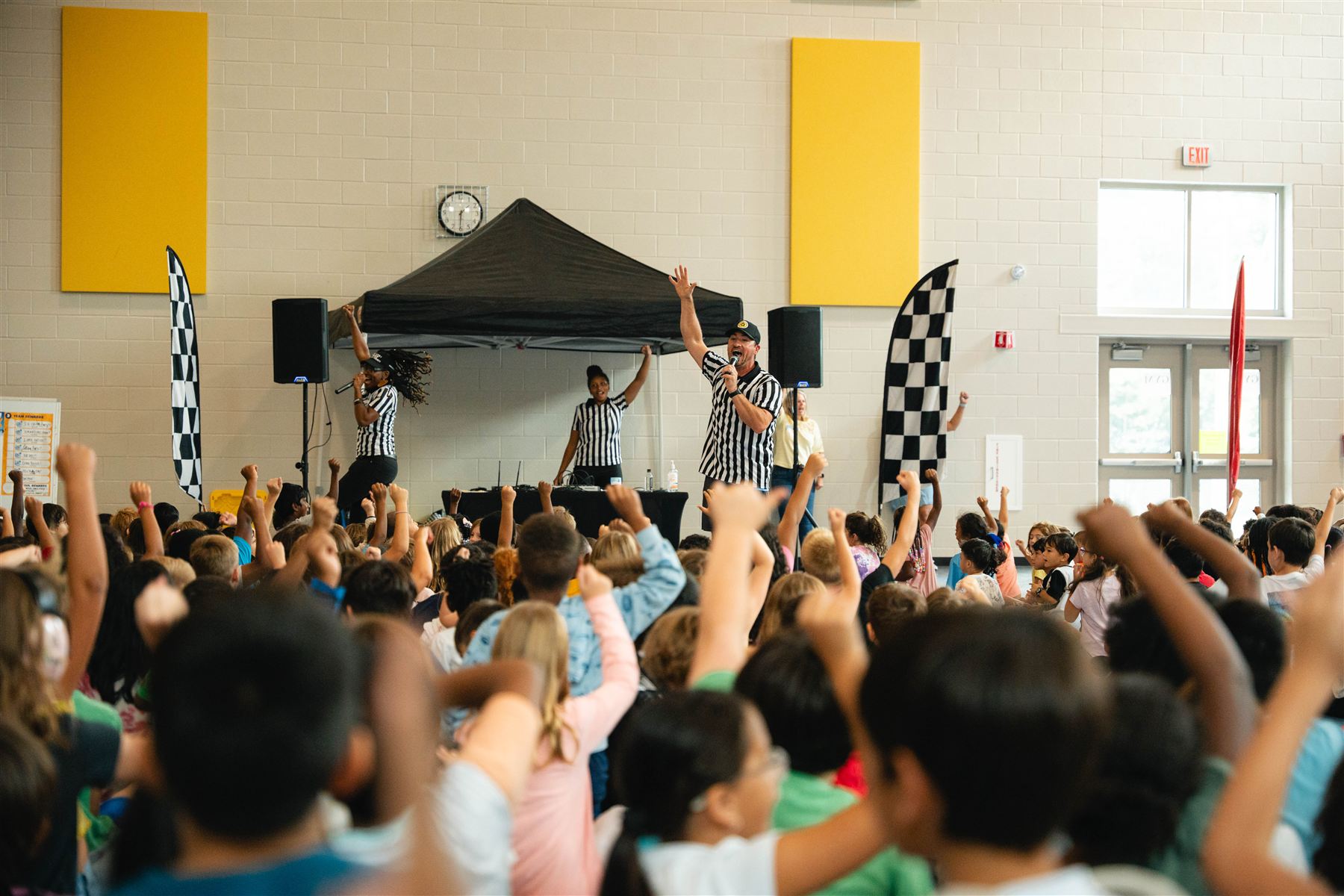 Students in a school gym raising their hands while two emcees lead from a stage